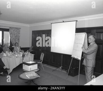 1980s, historisch, in einem Konferenzraum des Hotels, ein männlicher Trainer, der eine Präsentation mit Whiteboard und Overheadprojektor an die Unternehmensleiter, England, Großbritannien, vorstellte. Auf dem Whiteboard hat der Vortragende die Worte geschrieben, die Wahl; Change managen oder von IT verwaltet werden!“. Der Overheadprojektor oder OHP, wie er genannt wurde, war in dieser Zeit vor der Einführung des Diaprojektors und der Erstellung von „Audio-visuellen Diashows“ ein Standardinstrument für die Präsentation. Stockfoto