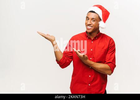 Fröhlicher und glücklicher junger Mann in rotem weihnachtsmann Hut zeigt mit Handflächen auf den leeren Raum, aufgeregt junge glücklich zu präsentieren Neuheit, Copy Space, Mockup, isoliert auf dem weißen Stockfoto