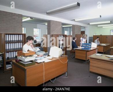 1989, historisches, männliches und weibliches Personal, das in einem Büro arbeitet, an Holzschreibtischen sitzt, in Tabletts sitzt, Maschinen und kleine Computer-Terminals des Tages hinzufügt. Stockfoto