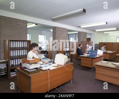 1989, historisches, männliches und weibliches Personal, das in einem Büro arbeitet, an Holzschreibtischen sitzt, in Tabletts sitzt, Maschinen und kleine Computer-Terminals des Tages hinzufügt. Stockfoto