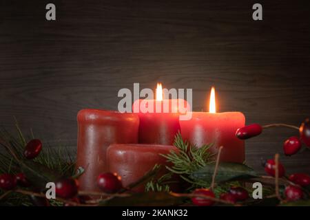 Adventskranz mit roten Kerzen mit zwei Kerzen, die für die zweite Adventwoche mit immergrünen Ästen und roten Beeren auf einem dunklen Raum mit Kopierfläche angezündet werden Stockfoto