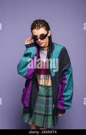 Junge Frau in Retro-Sportjacke mit Sonnenbrille auf violettem Hintergrund, Stockbild Stockfoto