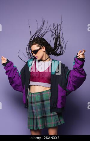 Stilvolle Frau in vintage Sportjacke und Sonnenbrille posiert auf lila Hintergrund, Stock Bild Stockfoto