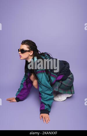 Frau mit Dreadlocks posiert in Vintage-Jacke und Sonnenbrille auf lila Hintergrund, Stockbild Stockfoto