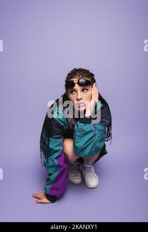 Frau mit geflochtenen Dreadlocks und Sonnenbrille sitzt auf Geisterbeinen in Neunzigern-Stil Jacke auf lila Hintergrund, Stockbild Stockfoto