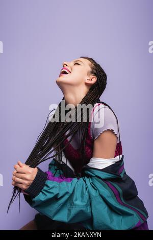 Aufgeregt Frau im Stil der neunziger Jahre Outfit mit geflochtenen Dreadlocks und Lachen isoliert auf lila, Stock Bild Stockfoto