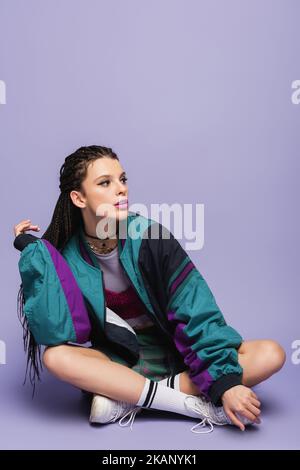 Frau mit Zöpfen sitzt in Neunzigern Stil Jacke und Blick weg auf lila Hintergrund, Stock Bild Stockfoto