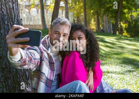 Ein lächelndes und glückliches Paar, das auf dem Gras im Park sitzt und ein Foto mit dem Handy macht Stockfoto