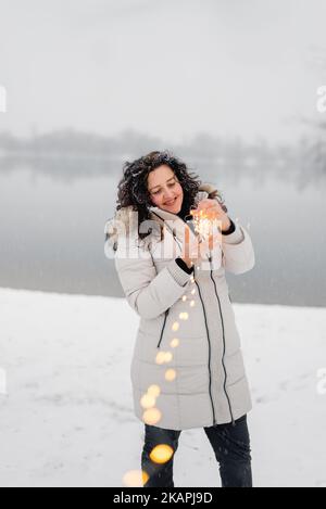 Weichzeichnerfokus. Ein wunderschönes Mädchen mit langen Locken hält an frostigen Tagen gelbe Girlande in den Händen. Frau, die unter Schnee und Schneeflocken kreist und das Leben im Freien genießt. Frohe Weihnachten Stockfoto