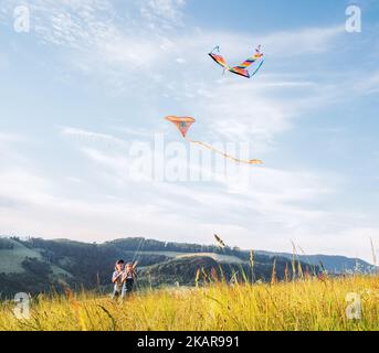 Lächelnde Schwester mit startenden Bruder mit bunten Drachen - beliebtes Outdoor-Spielzeug auf der Wiese mit hohen Grashügeln. Glückliche Kindheitsmomente oder Outdoor-tim Stockfoto