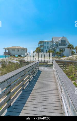 Destin, Florida Beach - Blick auf Strandhäuser von einer Holzpromenade aus. Es gibt Gräser in der Nähe der Promenade und Strandhäuser gegen den blauen Himmel. Stockfoto