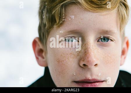 Blauäugiger Junge mit Sommersprossen und Schneeflocken im Gesicht Stockfoto