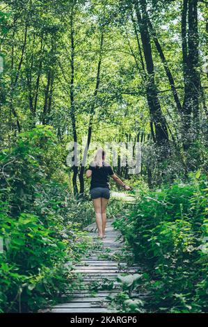 Frau geht auf alten Holzweg in grünen Walddickichten Stockfoto