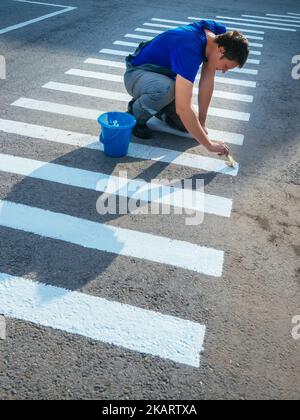 Straßendienstmitarbeiter sitzt auf seinem Schoß und malt gestreifte Fußgängerübergänge mit Pinsel auf Asphalt. Markierungen für Fußgänger auf der Straße. Sicherer Übergang für Menschen. Echter Workflow... Stockfoto