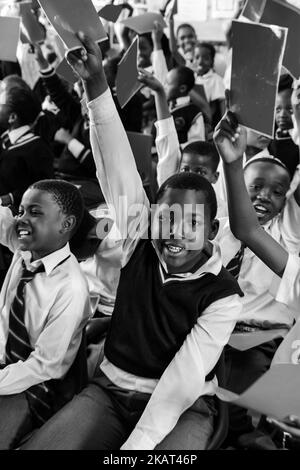 Eine Graustufenaufnahme einer Gruppe afrikanischer Kinder, die in einer Reihe sitzen und im Klassenzimmer der Grundschule Papiere aufziehen Stockfoto