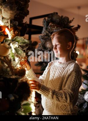Fröhliches und fröhliches kaukasisches Kind in der Nähe des Neujahrsbaums, das es mit Kugeln und einem LED-Blitz schmückte. Rotschopf kleiner Junge Dekoration Weihnachten 2023 Baum mit Lächeln. Weihnachtskonzept. Hochwertige Bilder Stockfoto