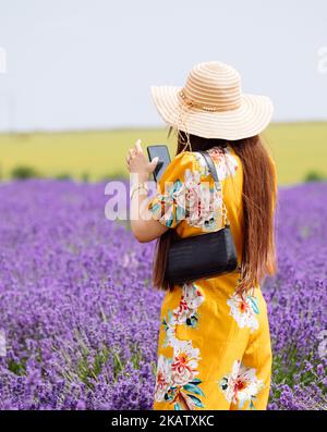 Eine junge asiatische Frau in einem gelben Jump-Anzug und einem breit gerömmten Strohhut, die ein Selfie in den Cotswold Lavendelfeldern bei Snowshill in Worcestershire gemacht hat. Stockfoto