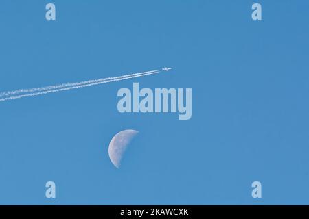 Hochfliegender Passagierjet-Liner mit nachliegendem Kondensstreifen, der einen Halbmond vor einem klaren, blauen Himmel passiert Stockfoto