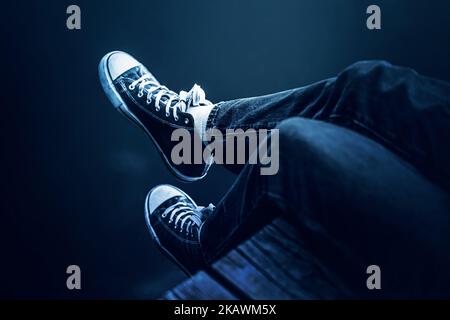 Beine in Turnschuhen und blaue Jeans eines Menschen, der auf einem hölzernen Pier über dem dunklen Wasser des Sees sitzt. Lifestyle und Erholung am Wasser. Stockfoto