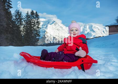 Das kleine Mädchen sitzt auf einem Schlitten und zeigt sich über die Berge, die bergab fahren Stockfoto