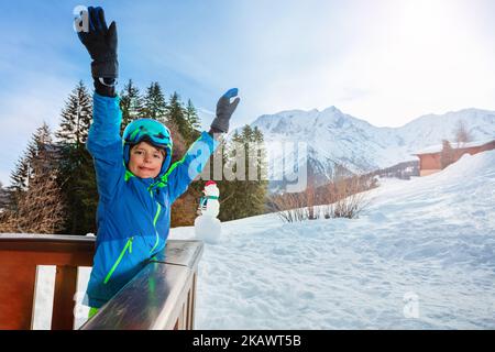 Kleiner Junge im Skioutfit steht auf dem Balkon und hebt die Hände Stockfoto