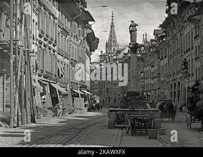 Eine Straßenszene auf der Marktgasse in der ‘Altstadt’, Bern, Schweiz, c. 1900. Der Anna-Seiler-Brunnen ist ein Springbrunnen, dessen Mittelpunkt der Brunnen ist. Geschäfte und Händler säumen die Straße und das Pferd und der Wagen bilden einen wichtigen Verkehrsträger, aber auch Straßenbahnschienen und die damit verbundene Überkopfverdrahtung sind sichtbar. Der Brunnen ist Anna Seiler gewidmet, der Gründerin des ersten Spitals in Bern. Anna Seiler gründete in ihrem Haus ein Krankenhaus. Dieses Bild stammt aus einem alten Glasnegativ – einem viktorianischen Vintage-Foto. Stockfoto