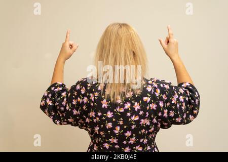 Die lateinamerikanische Blondine in einem dunklen Kleid, von hinten gedreht, zeigt mit ihren Zeigefingern nach oben, isoliert auf einem beigen Hintergrund. Stockfoto