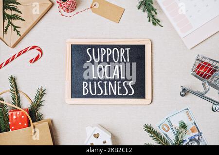 Support Local Business Text auf Tafel geschrieben. Flat Lay mit Unterstützung Local Business Zitat, Weihnachtsdekoration und Einkaufstasche, Geschenkboxen und Stockfoto