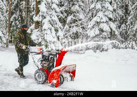 Im Winter reinigt ein Mann mit einem Schneepflug im Wald vor dem Hintergrund eines verschneiten Waldes die Straße vom Schnee. Stockfoto