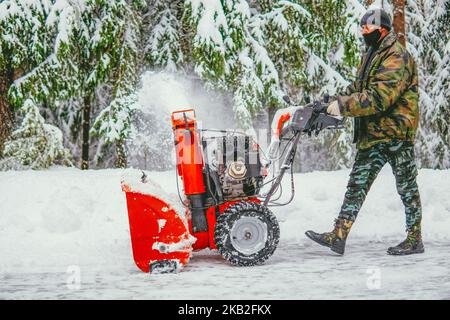 Im Winter reinigt ein Mann mit einem Schneepflug im Wald vor dem Hintergrund eines verschneiten Waldes die Straße vom Schnee. Stockfoto