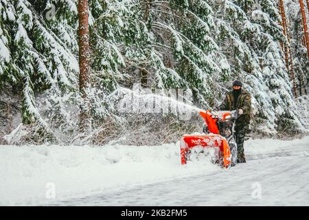 Im Winter reinigt ein Mann mit einem Schneepflug im Wald vor dem Hintergrund eines verschneiten Waldes die Straße vom Schnee. Stockfoto