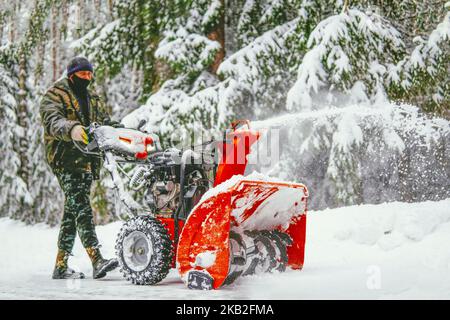 Ein Mann entfernt Schnee mit einem Schneefräsen auf dem Hintergrund eines verschneiten Waldes Stockfoto