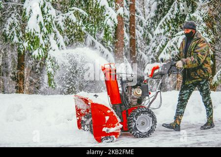 Ein Mann entfernt Schnee mit einem Schneefräsen auf dem Hintergrund eines verschneiten Waldes Stockfoto