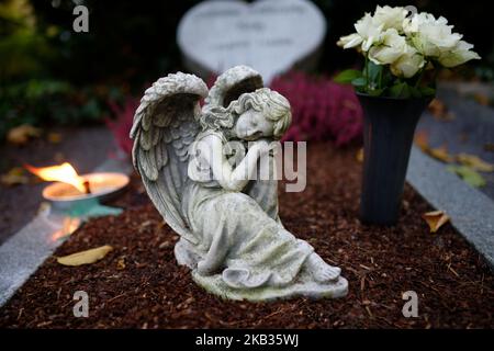 Kleine Engelsfigur mit Flügeln auf einem Grab mit Blumen und brennender Kerze in der Dämmerung Stockfoto