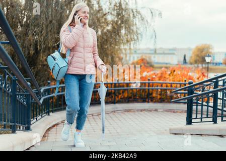 Ein blondes Mädchen spricht am Telefon und geht vor dem Hintergrund herbstlicher Blätter durch die Stadt und lacht an einem sonnigen Tag. Stockfoto