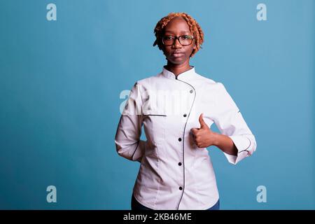Seriöse afroamerikanische Köchin, die mit Daumen nach oben steht und die Kamera anschaut. Professionelle Köchin in Uniform zeigt Daumen nach oben Geste der Zustimmung vor blauem Hintergrund. Stockfoto