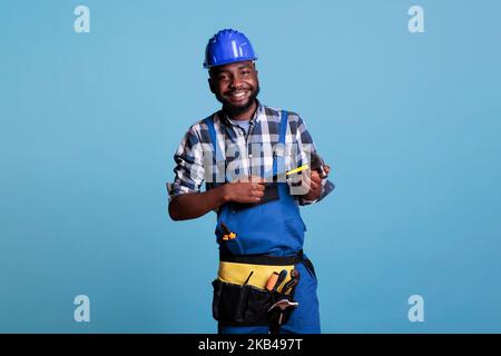 Glücklicher Bauarbeiter, der vor blauem, isoliertem Hintergrund optimistisch lächelt. Studiofoto eines afroamerikanischen Mannes mit einem Hut und einem Werkzeuggürtel, der den Hammer mit beiden Händen hält. Stockfoto