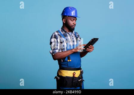 Porträt eines Baumeisters, der mit einem digitalen Tablet arbeitet und Helmschutz trägt. Bauprofi mit einem Touchscreen-Gerät während seiner Arbeitsschicht im Studio vor blauem Hintergrund aufgenommen. Stockfoto