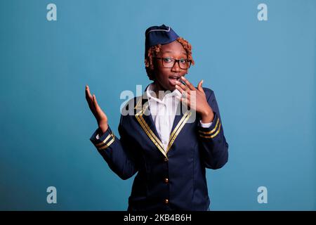 afroamerikanische Stewardess, die Überraschung, Erstaunen ausrief. Flugcrew-Frauenstudioportrait mit blauem Hintergrund, lockiges, afro Haar in Ehrfurcht, beeindruckt. Stockfoto