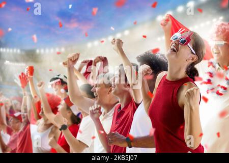 Fußballfan im Stadion in rot-weißem Hemd. Fröhliche Fans auf dem Fußballfeld, die das siegreiche Mannschaftsspiel beobachten. Gruppe von Unterstützern mit Flagge Stockfoto