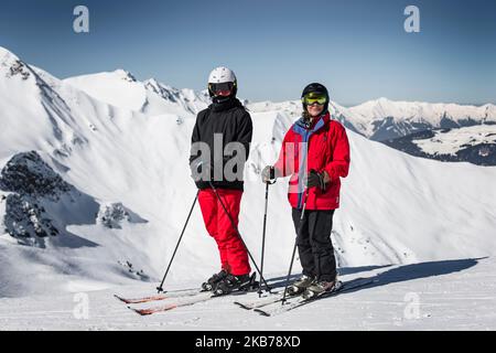 Zwei Skifahrer eine Frau ein Mann hält am Rand des Berges das Foto, hohe Berge der Alpen im Hintergrund auf einem sonnigen Tag Helme und Brillen auf roten und schwarzen Outfits Stockfoto