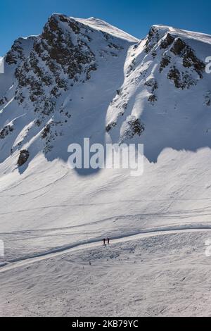 Zwei Skifahrer, die von der Größe der Berge rund um die Piste umgeben sind, fahren zwischen den Bergen blaue Himmelsschatten ab Stockfoto