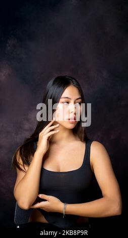 Multirassische Teen mit langen schönen Haaren sitzend mit Armen in Tanzpose mit Blick weg von der Kamera mit Copy Space über ihr Stockfoto