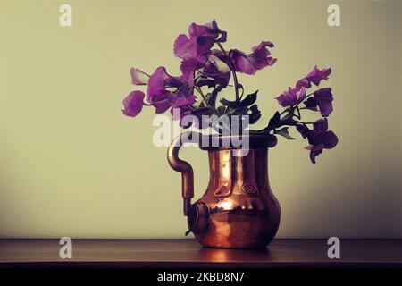 Kreative und Stillleben-Illustration zartsüßer Erbsenblüten in einem glänzenden Kupfertopf auf einem Tisch. Stockfoto