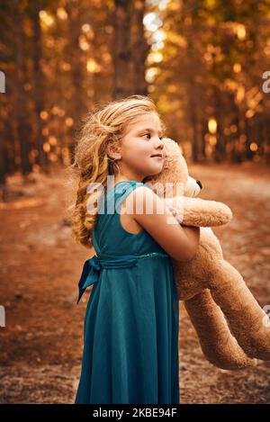 Mein Teddy geht wohin ich gehe. Ein glückliches kleines Mädchen hält ihren Teddybär und blickt in die Ferne, während es draußen im Wald steht. Stockfoto