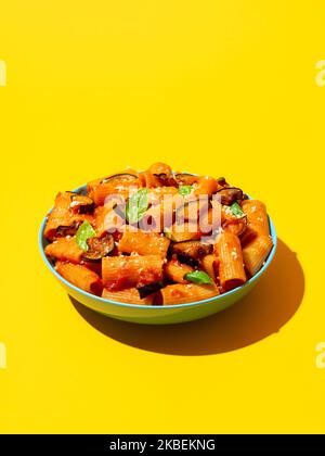 Pasta Alla Norma Schale minimalistisch auf einem gelben Tisch in hellem Licht. Köstliches italienisches Gericht, Pasta Rigatoni mit Tomatensauce und frittierter Aubergine. Stockfoto