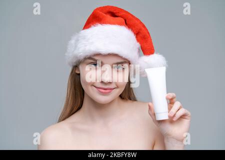 Portrait junge, gesunde, gepflegte Frau im weihnachtsmütze mit weißem Röhrenmockup in der Hand, Hautpflegekonzept, Nahaufnahme auf grauem Hintergrund. Beauty Kosmetik und Kosmetik im Winter Stockfoto