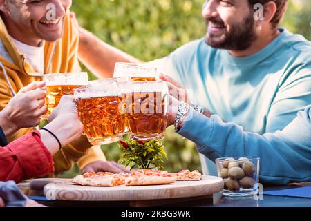 Fröhliche Gruppe junger Menschen, die Stimmungsgläser über einer Pizza in Scheiben klirren - Millennial-Freunde feiern das Beigehen von Bierkrügen beim Picknick im Freien in einem Su Stockfoto