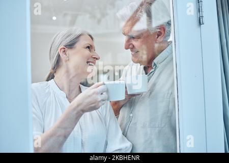 Liebe, Kaffee und Fenster mit einem älteren Paar entspannen, lachen und binden, während sie gemütlich, glücklich und trinken Kaffee in ihrem Haus. Glas, Tee und ältere Menschen Stockfoto