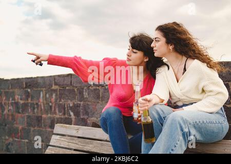 Zwei junge Freundinnen zeigen weg und trinken Bier aus Flaschen und sitzen auf einer Bank im Freien - Menschen, Essen und Trinken Lifestyle-Konzept Stockfoto